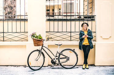 Portrait of a young beautiful woman with bicycle and smartphone standing against a building. Sunny spring town. - HPIF25307