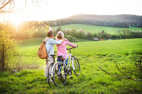 Beautiful senior couple outside in spring nature, standing with bicycles on grassland. Rear view. - HPIF25394