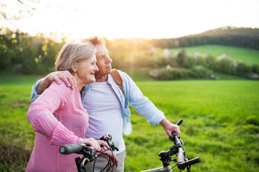 Beautiful senior couple outside in spring nature, standing with bicycles on grassland. - HPIF25395