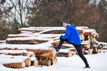 Ein älterer Mann, der sich vor dem Lauf in der winterlichen Natur am Holzstapel die Beine ausstreckt. Platz kopieren. - HPIF25439