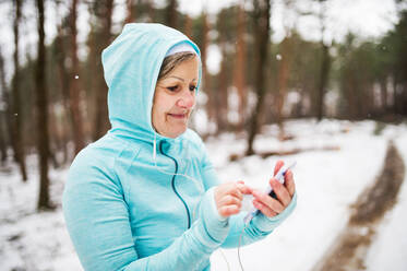 Ältere Läuferin mit Kopfhörern und Smartphone in winterlicher Natur stehend, Musik hörend und sich ausruhend, Rückansicht. - HPIF25445