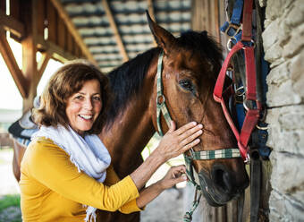 A senior woman with a brown horse standing by a stable. - HPIF25493