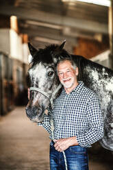 Ein glücklicher älterer Mann steht in der Nähe eines Pferdes in einem Stall und hält es. - HPIF25515