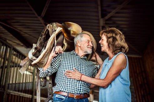 Ein glückliches älteres Paar, das in einem Stall einen Pferdesattel auf den Schultern trägt. - HPIF25523