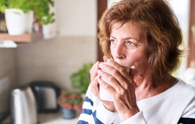 Ältere Frau in der Küche. Eine alte Frau im Haus, die eine Tasse Kaffee in der Hand hält und trinkt. - HPIF25562