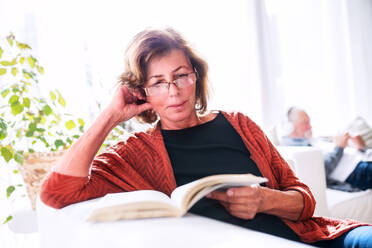 Happy senior couple relaxing at home. A woman reading, and unrecognizable man in the background. - HPIF25577