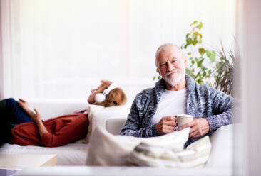 Ein glückliches älteres Ehepaar sitzt zu Hause auf dem Sofa und entspannt sich, eine Frau hört Musik und ein Mann hält eine Tasse Kaffee. - HPIF25580