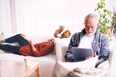 Ein glückliches älteres Ehepaar sitzt auf dem Sofa und entspannt sich zu Hause. Eine Frau hört Musik und ein Mann benutzt ein Tablet. - HPIF25581