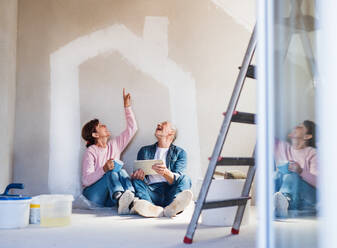 Happy senior couple painting walls in new home, using tablet. Relocation concept. - HPIF25630