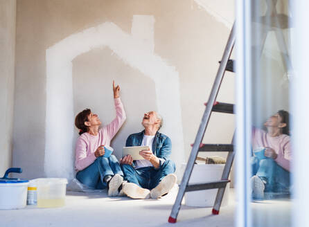 Glückliches älteres Ehepaar, das mit Hilfe eines Tablets die Wände in seiner neuen Wohnung streicht. Konzept des Umzugs. - HPIF25630