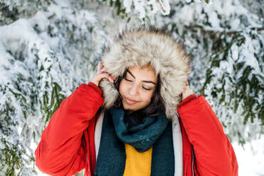 Porträt einer jungen Frau, die im Freien in einem verschneiten Winterwald steht und eine Pelzkapuze trägt. - HPIF25817