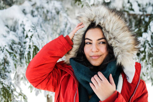 Porträt einer jungen Frau, die im Freien in einem verschneiten Winterwald steht und eine Pelzkapuze trägt. - HPIF25818