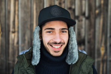 Front view portrait of young man standing against wooden background outdoors in winter. - HPIF25823