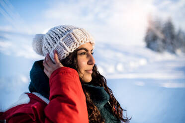 A front view portrait of young woman standing outdoors in snowy winter forest, wearing woollen hat. - HPIF25828