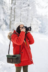 Eine junge Frau mit Kamera steht im Freien im Schnee im Winterwald und macht Fotos. - HPIF25846