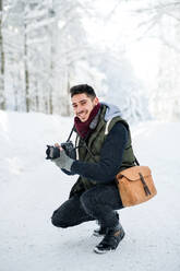 Ein junger Mann mit Kamera im Freien im Schnee im Winterwald, der in die Kamera schaut. - HPIF25848