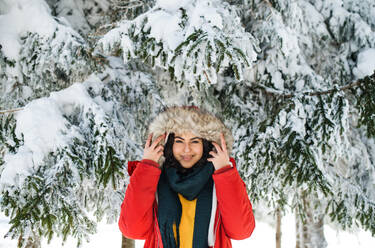 Porträt einer jungen Frau, die im Freien in einem verschneiten Winterwald steht und eine Pelzkapuze trägt. - HPIF25877