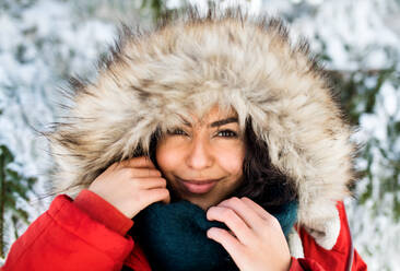 Porträt einer jungen Frau, die im Freien in einem verschneiten Winterwald steht und eine Pelzkapuze trägt. - HPIF25878