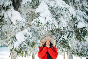 Porträt einer jungen Frau, die im Freien in einem verschneiten Winterwald steht und eine Pelzkapuze trägt. - HPIF25879