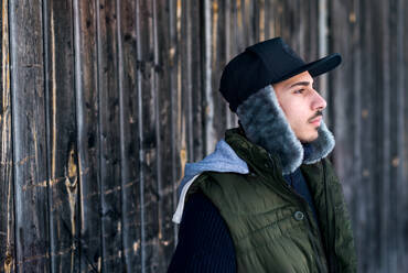 Side view portrait of young man standing against wooden background outdoors in winter. - HPIF25898