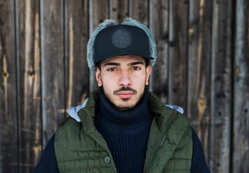 Front view portrait of young man with cap standing against wooden background outdoors in winter, close-up. - HPIF25899