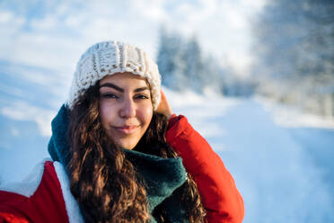 A front view portrait of young woman standing outdoors in snowy winter forest, wearing woollen hat. - HPIF25902