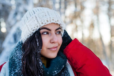 A front view portrait of young woman standing outdoors in snowy winter forest, wearing woollen hat. - HPIF25916