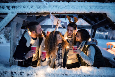 A group of young friends outdoors in snow in winter at night, holding drinks. - HPIF25940