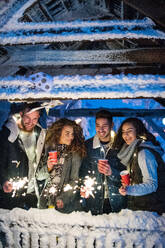 A group of young friends outdoors in snow in winter at night, holding sparklers. - HPIF25944