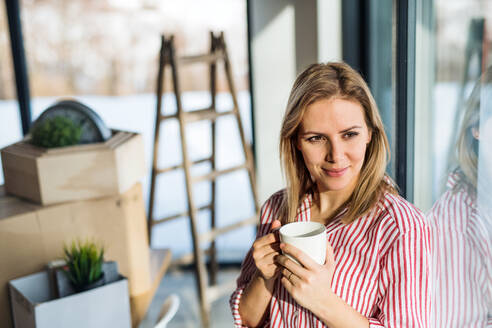 Eine glückliche junge Frau zieht in ein neues Haus ein, lehnt sich an ein Fenster und trinkt Kaffee. - HPIF26022