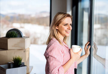 Eine glückliche junge Frau zieht in ein neues Haus ein, lehnt sich an ein Fenster und trinkt Kaffee. - HPIF26023