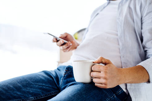 Ein unkenntlicher Mann mit Smartphone und Kaffee sitzt in einem Haus und schreibt eine SMS. - HPIF26045