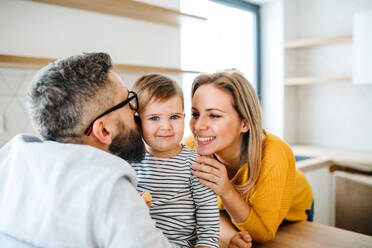 A portrait of young happy family with a toddler girl indoors in kitchen, kissing. - HPIF26074