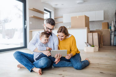 A young family with a toddler girl sitting barefooton the floor when moving in new home, using tablet. - HPIF26082