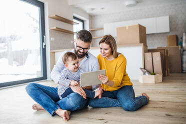 A young family with a toddler girl sitting barefooton the floor when moving in new home, using tablet. - HPIF26083