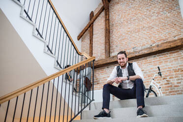 Ein junger Geschäftsmann sitzt auf einer Treppe in einem Bürogebäude und schaut in die Kamera. - HPIF26153