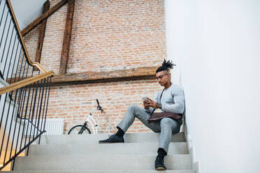 Ein junger gemischtrassiger Geschäftsmann mit Telefon sitzt auf einer Treppe in einem Bürogebäude. - HPIF26160