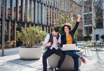 Young businesspeople with laptop outdoors in courtyard, expressing excitement. Start-up concept. - HPIF26194
