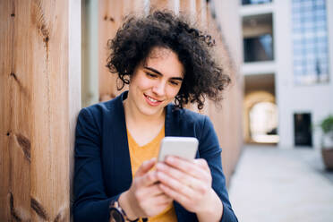 A happy young business woman with smartphone standing outdoors, text messaging. Start-up concept. - HPIF26214