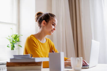 Eine junge, glückliche Studentin, die zu Hause am Tisch sitzt und einen Laptop benutzt, um zu lernen. - HPIF26235