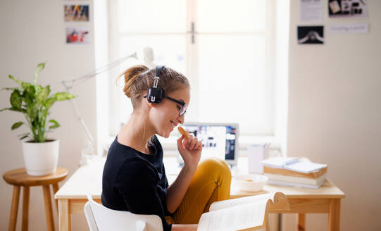 Eine junge, glückliche Studentin sitzt zu Hause am Tisch und benutzt Kopfhörer und Laptop beim Lernen. - HPIF26259