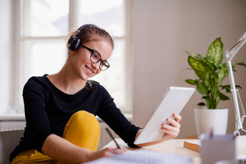 Eine junge, glückliche Studentin sitzt zu Hause am Tisch und benutzt beim Lernen Kopfhörer und Tablet. - HPIF26262