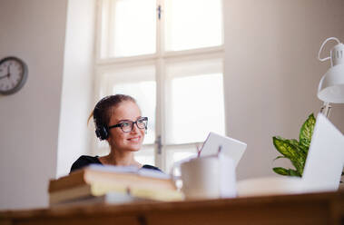 A young happy college female student sitting at the table at home, using headphones and tablet when studying. - HPIF26264