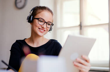 Eine junge, glückliche Studentin sitzt zu Hause am Tisch und benutzt beim Lernen Kopfhörer und Tablet. - HPIF26265