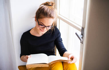 Eine junge glückliche Studentin mit einem Buch, die zu Hause auf der Fensterbank sitzt und studiert. - HPIF26272