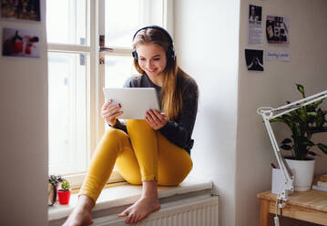 Eine junge, glückliche Studentin, die zu Hause auf der Fensterbank sitzt und beim Lernen Tablet und Kopfhörer benutzt. - HPIF26281