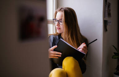 A young happy college female student with an exercise book sitting on window sill at home, studying. - HPIF26283