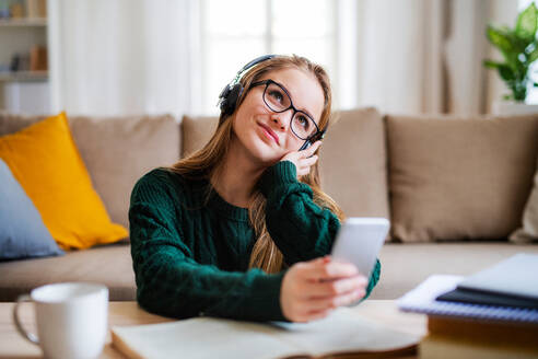 Eine junge, glückliche Studentin sitzt zu Hause am Tisch und benutzt beim Lernen Kopfhörer. - HPIF26291