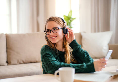 Eine junge, glückliche Studentin sitzt zu Hause am Tisch und benutzt beim Lernen Kopfhörer. - HPIF26292