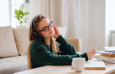 Eine junge, glückliche Studentin sitzt zu Hause am Tisch und benutzt beim Lernen Kopfhörer. - HPIF26293